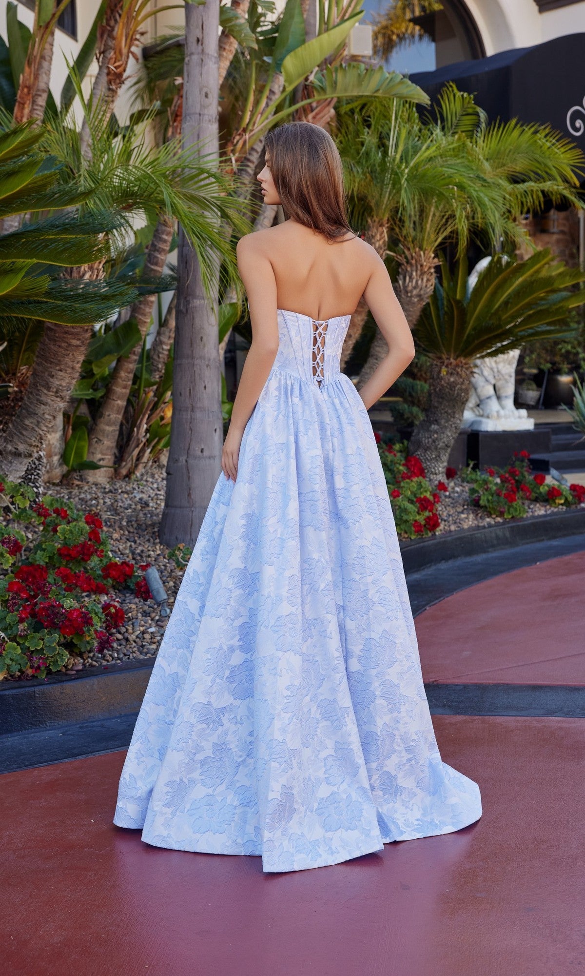 A woman stands outdoors with her back to the camera, wearing the Nox Anabel T1912 long prom dress in light blue floral jacquard with a lace-up back, surrounded by palm trees and red flowers.